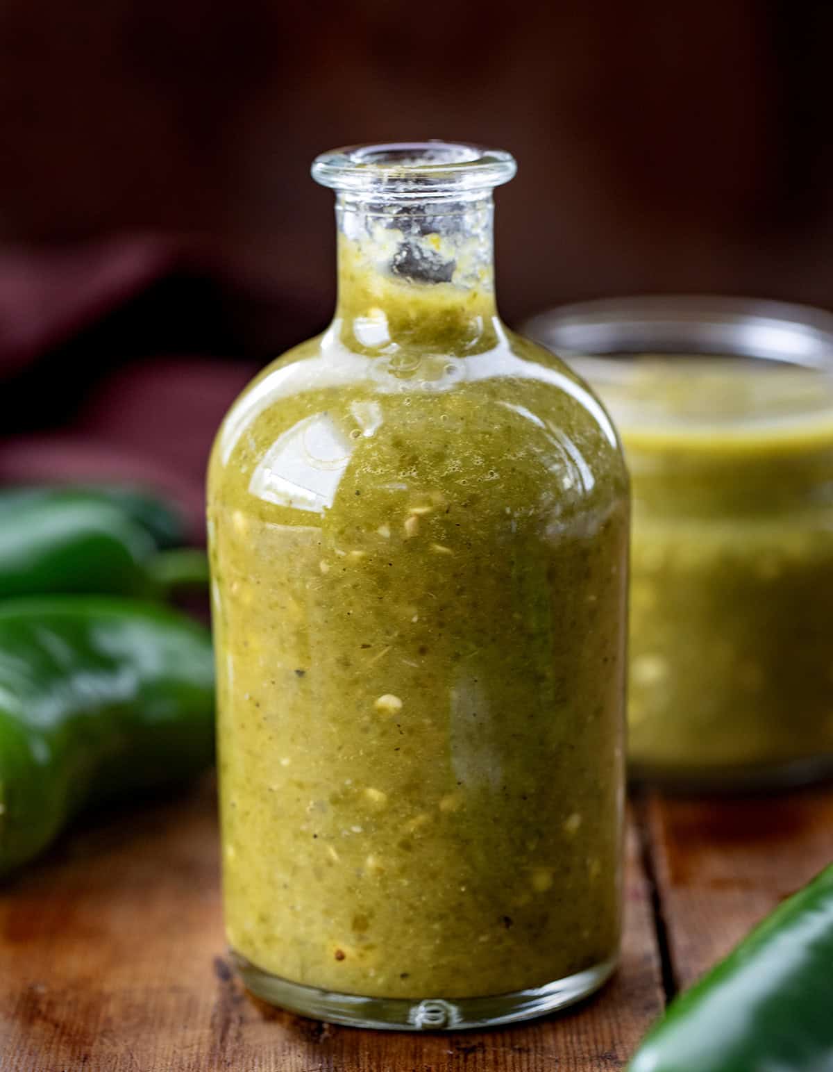 Jalapeno Hot Sauce in a jar on a wooden table with another jar in the background and fresh uncut jalapenos. 