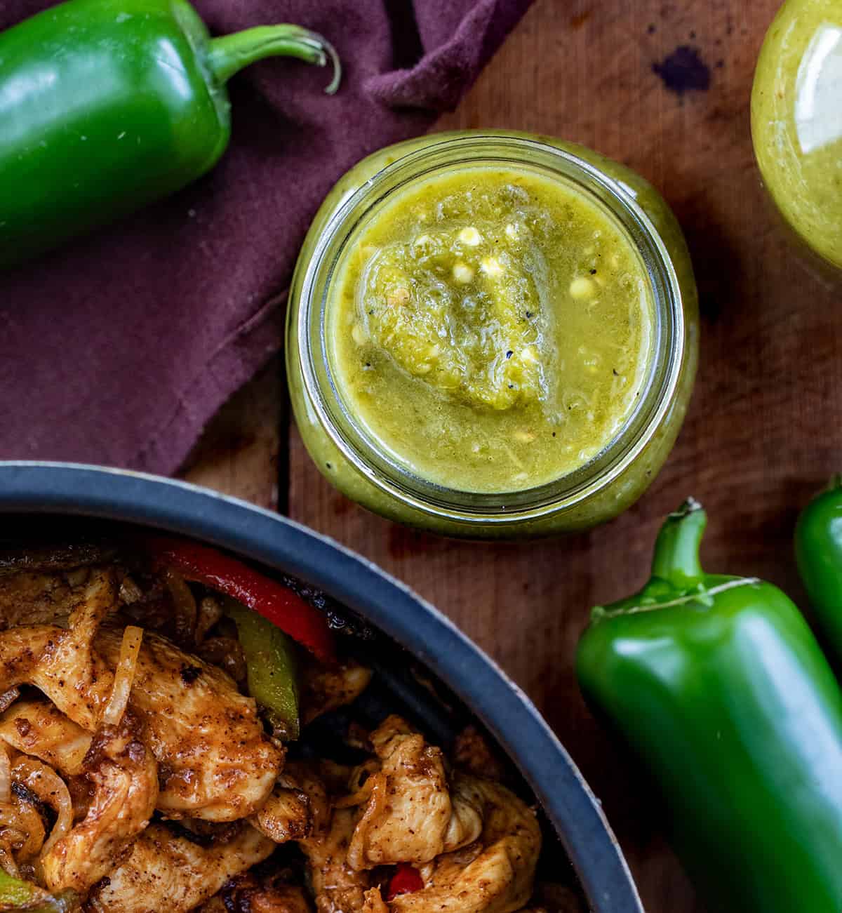 Jalapeno Hot Sauce in an open jar next to fajita chicken and jalapenos on a wooden table from overhead.