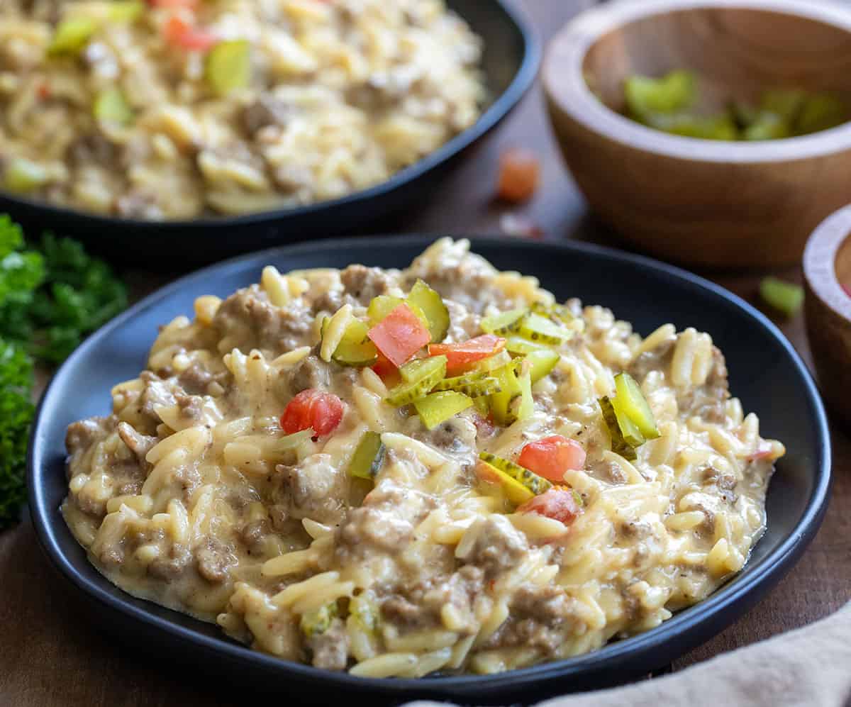 Plates of Cheeseburger Orzo on a wooden table topped with chopped pickle and chopped tomato.