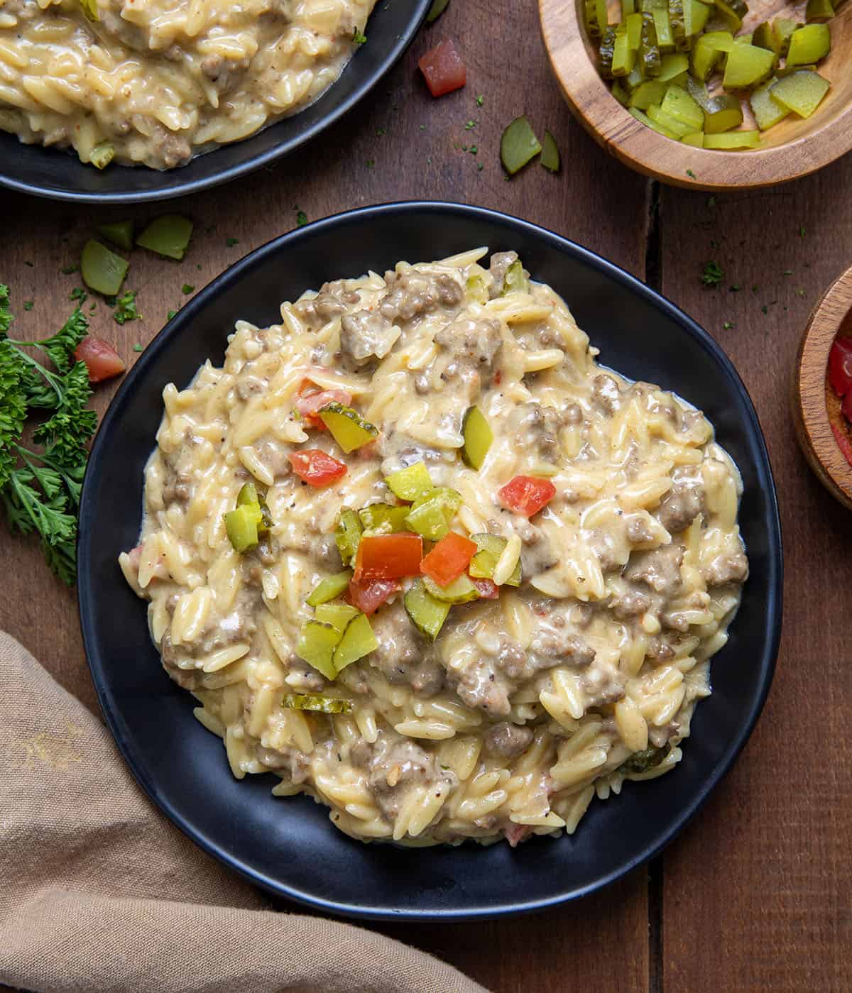 Plates of Cheeseburger Orzo on a wooden table with pickle and tomato from overhead.