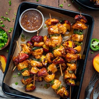 Jalapeno Peach Chicken Skewers in a tray on a wooden table surrounded by fresh peaches and diced jalapeno from overhead.
