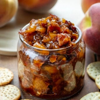 Jar of Peach Bacon Jam on a wooden table with crackers and fresh peaches.