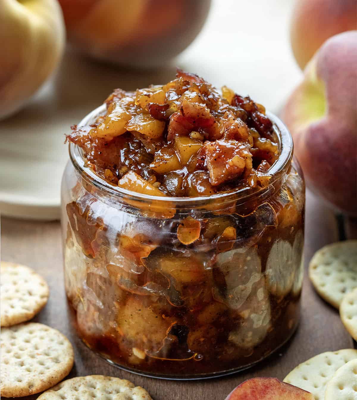 Jar of Peach Bacon Jam on a wooden table with crackers and fresh peaches. 