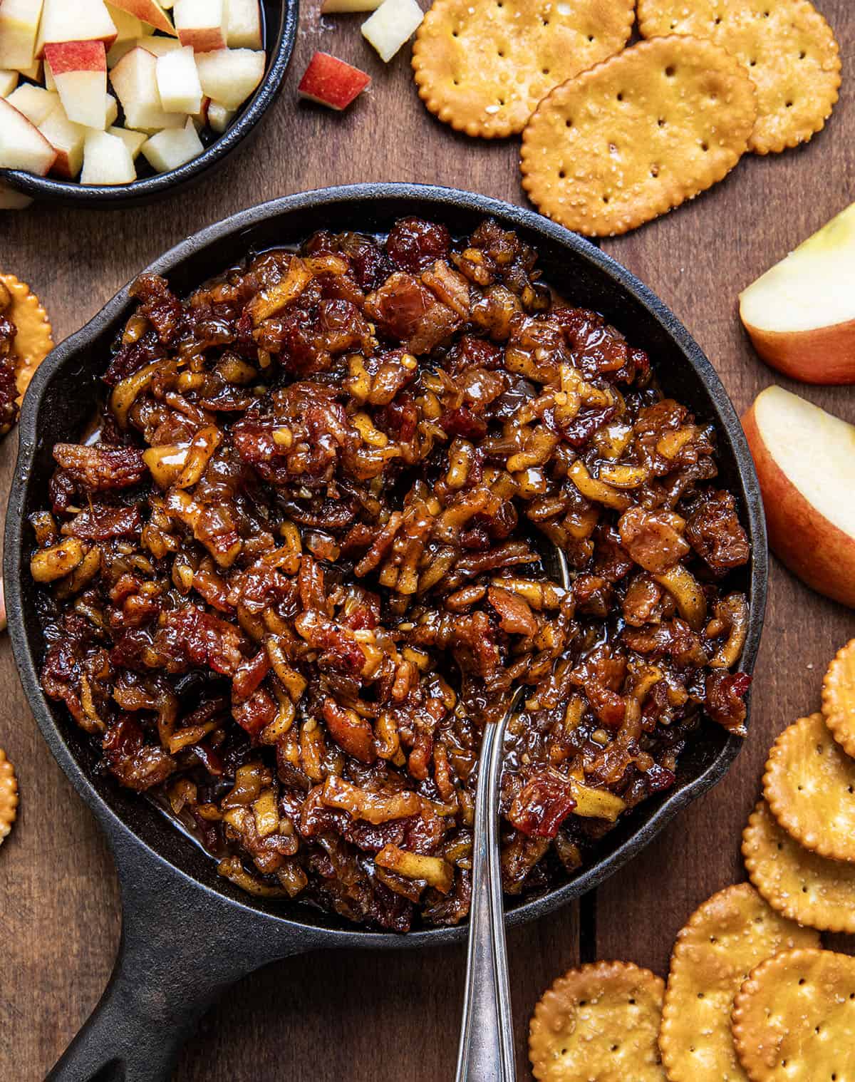 Mini Skillet of Apple Whiskey Bacon Jam surrounded by crackers and diced apples. 

