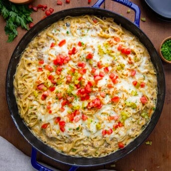 Skillet of Cheeseburger Tetrazzini on a wooden table from overhead surrounded by diced tomatoes, diced pickles, and a towel.