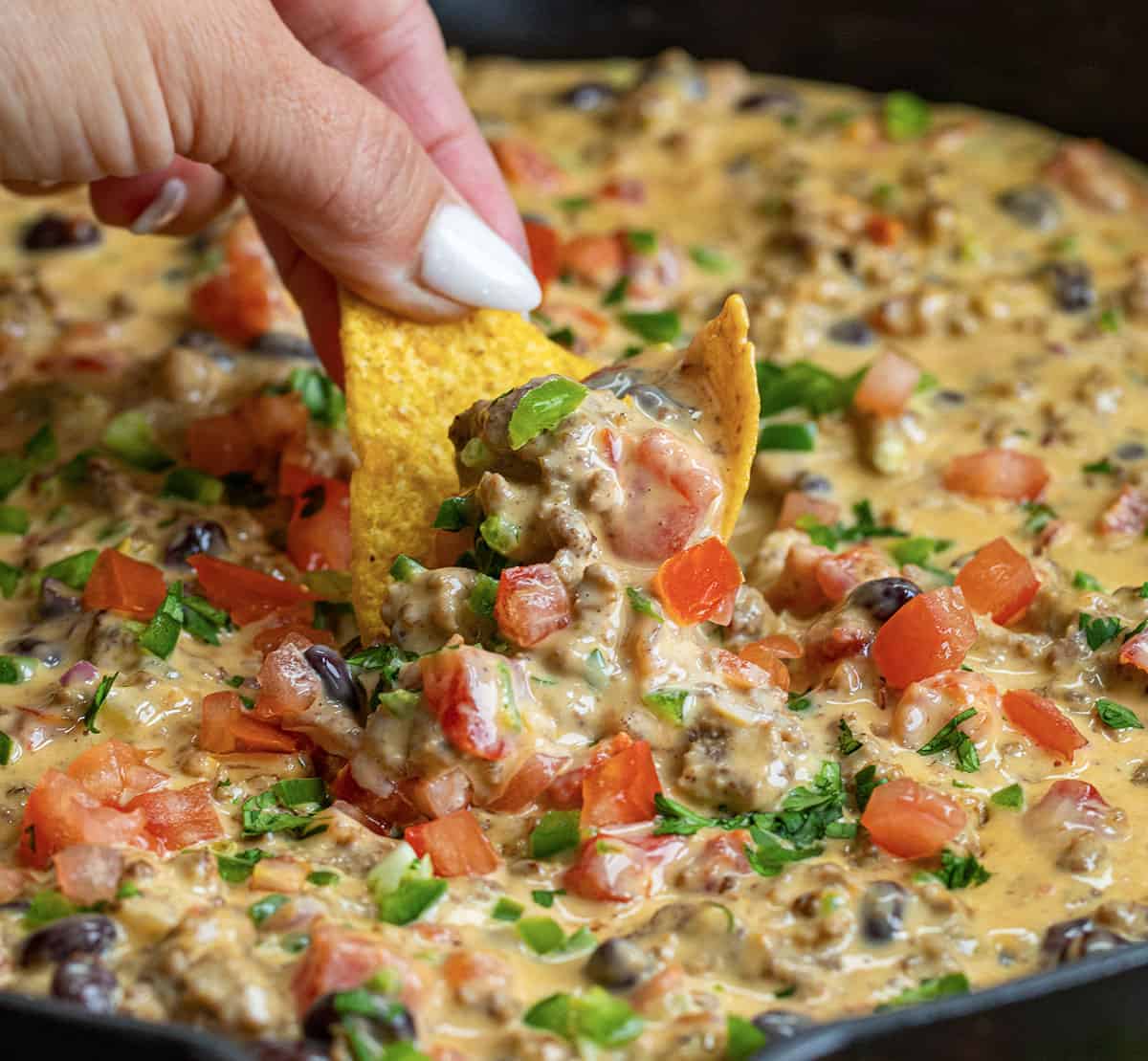 Dipping a chip into a skillet of Cowboy Queso topped with fresh tomato and jalapeno.