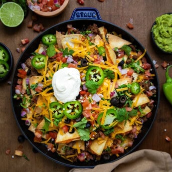 Skillet of Quesadilla Nachos on a wooden table loaded hight with guacamole, sour crea, cheese, jalapeno rounds, and olives from overhead.