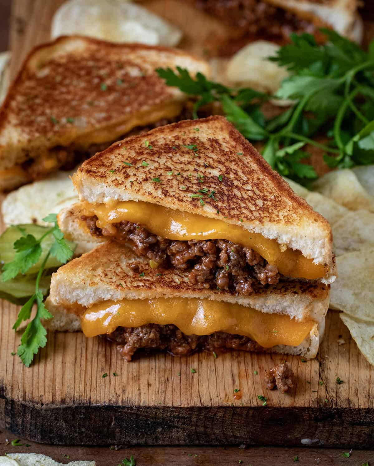 Sloppy Joe Grilled Cheese stacked on a cutting board with chips and more sandwiches in the background.