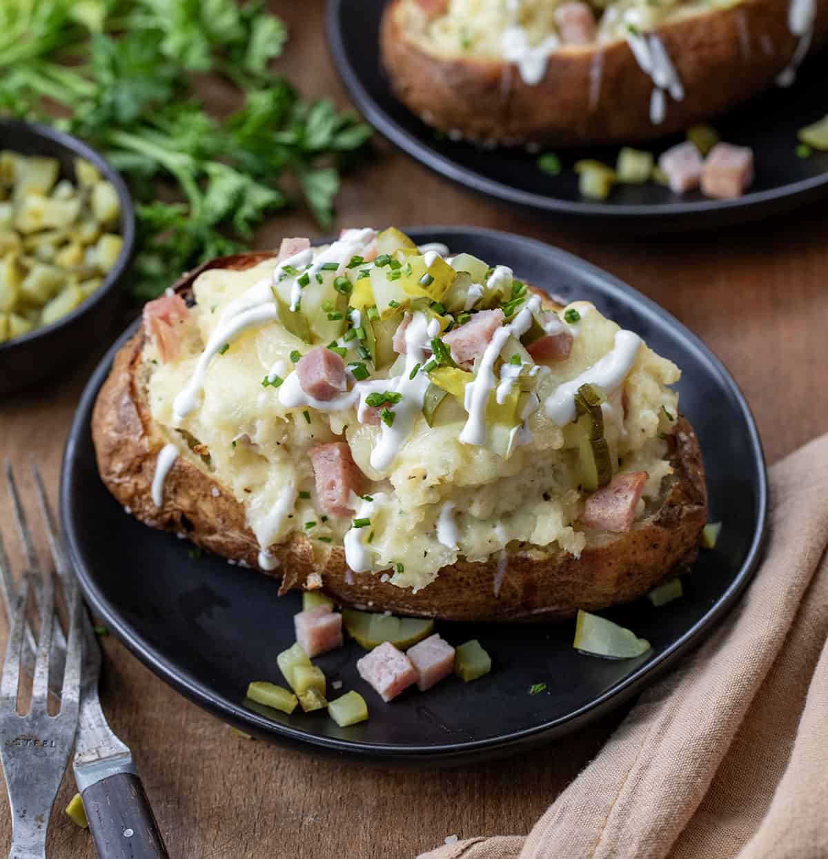 Dill Pickle and Ham Twice Baked Potatoes on black plates on a wooden table with a napkin and forks.