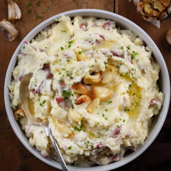 Bowl of Roasted Garlic Mashed Potatoes on a wooden table with a spoon in it.