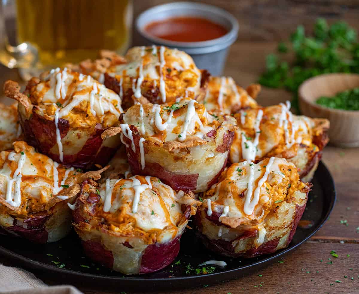 Buffalo Chicken Smashed Potato Cups on a skillet on a wooden table with buffalo sauce and beer in the background.