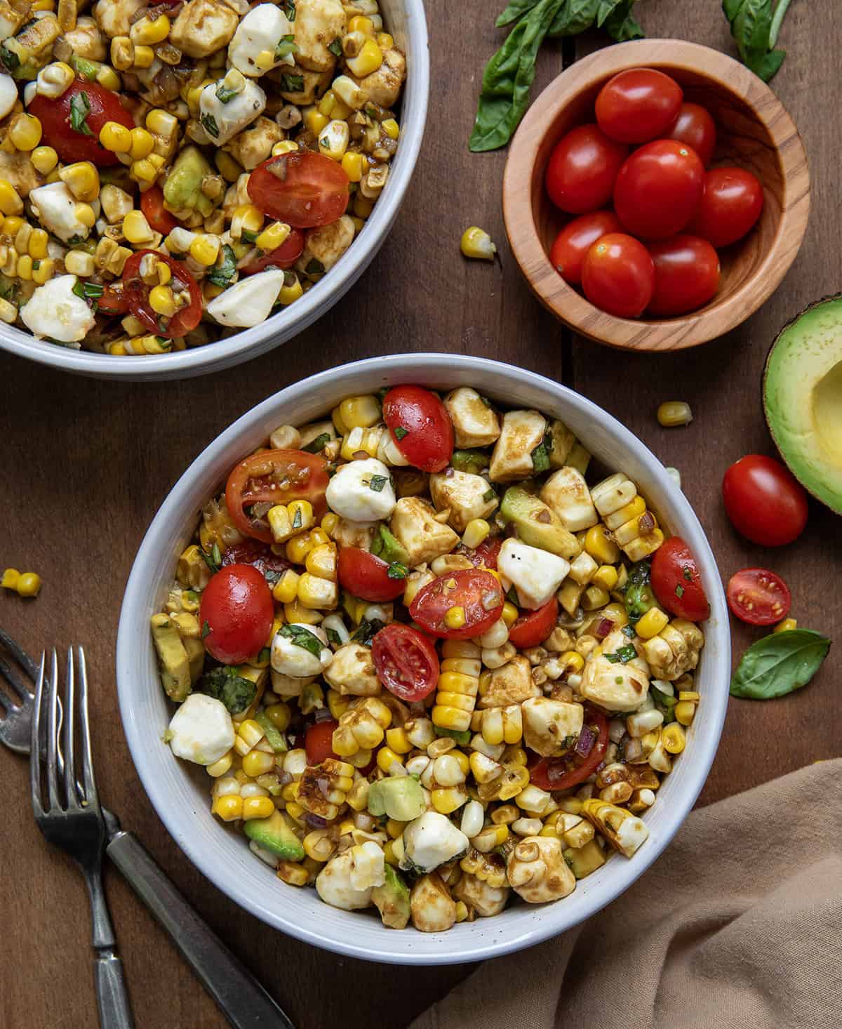 Bowls of Caprese Corn Salad on a wooden table with fresh tomatoes and avocado from overhead.