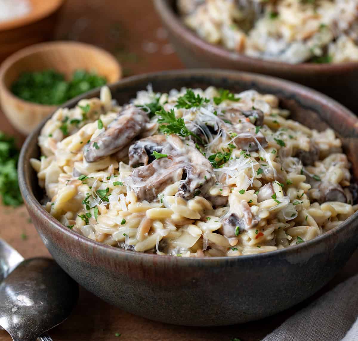 Bowls of Creamy Mushroom Orzo on a wooden table with a napkin and silverware around bowl.