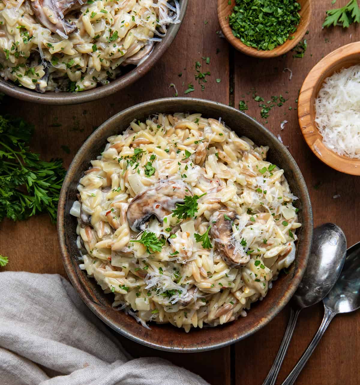 Bowls of Creamy Mushroom Orzo on a wooden table with parsley and freshly grated parmesan cheese from overhead.