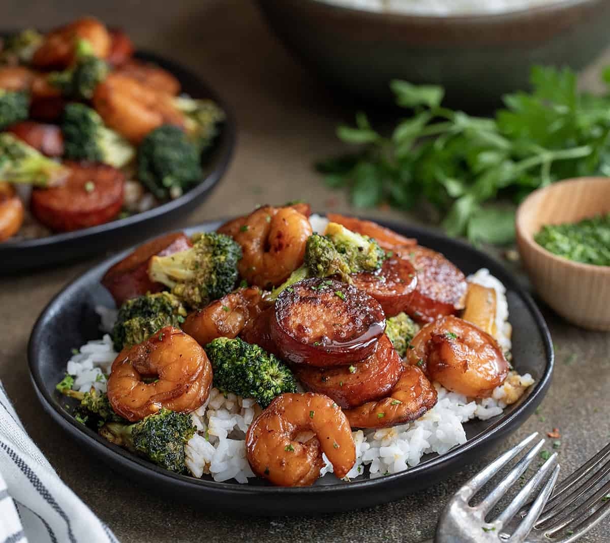 Black plate with rice and Honey Garlic Shrimp, Sausage, and Broccoli on a wooden table.