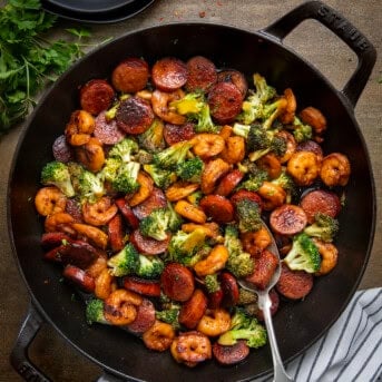 Skillet of Honey Garlic Shrimp, Sausage, and Broccoli with a spoon in it on a wooden table from overhead.