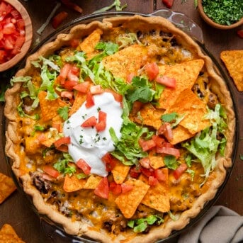 Whole Dorito Taco Pie on a wooden table with tomatoes, lettuce, sour cream, and doritos.