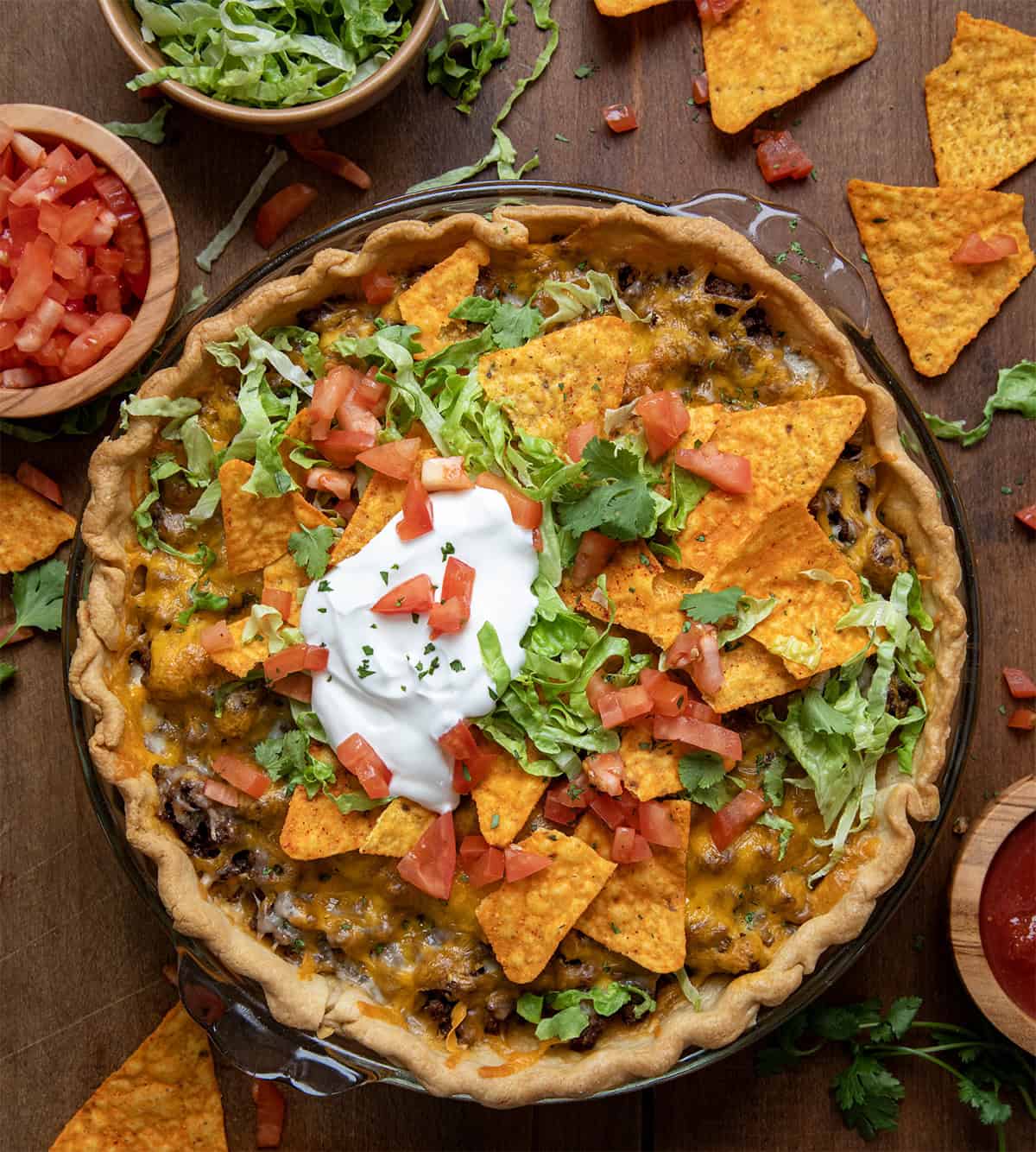 Whole Dorito Taco Pie on a wooden table with tomatoes, lettuce, sour cream, and doritos.