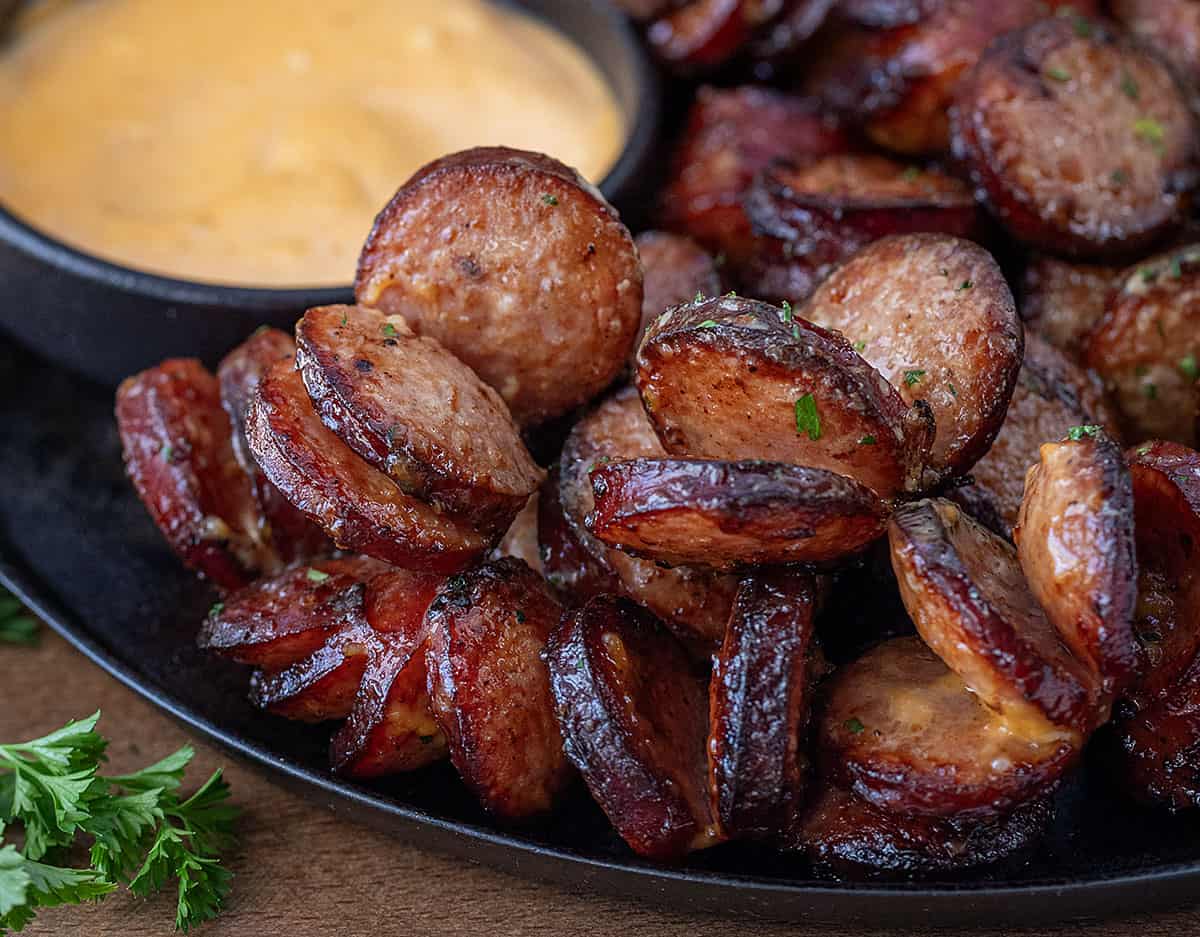 Very close up of Spicy Sriracha Hasselback Kielbasa Bites on a black skillet with sauce in background.