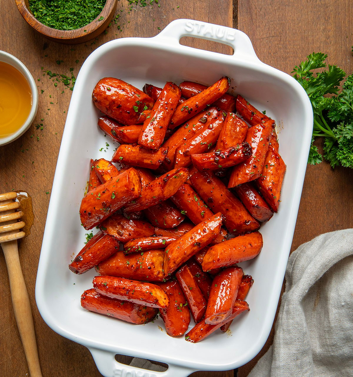 White pan of Honey Balsamic Roasted Carrots on a wooden table with honey and a towel from overhead.