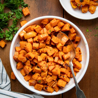 White Serving Bowl of Maple Chipotle Roasted Sweet Potatoes with a spoon in it on a wooden table from overhead.