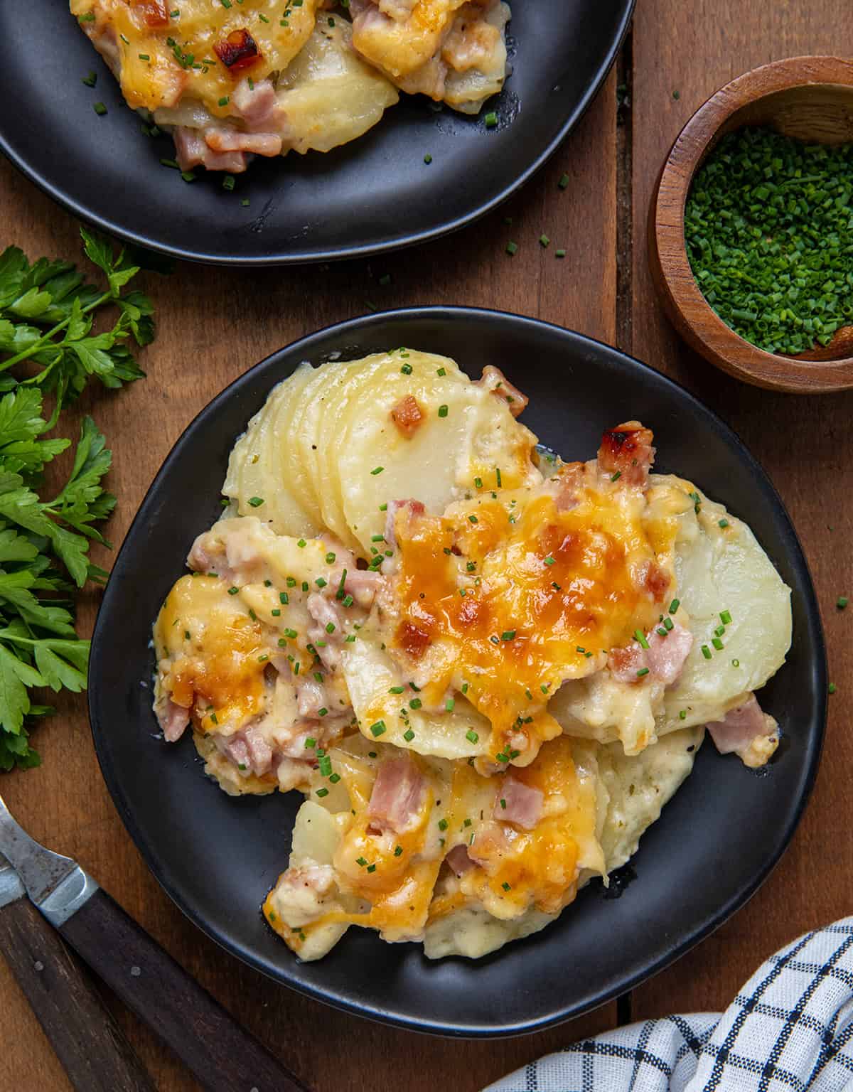 Black plates of Ranch Scalloped Potatoes and Ham on a wooden table from overhead.