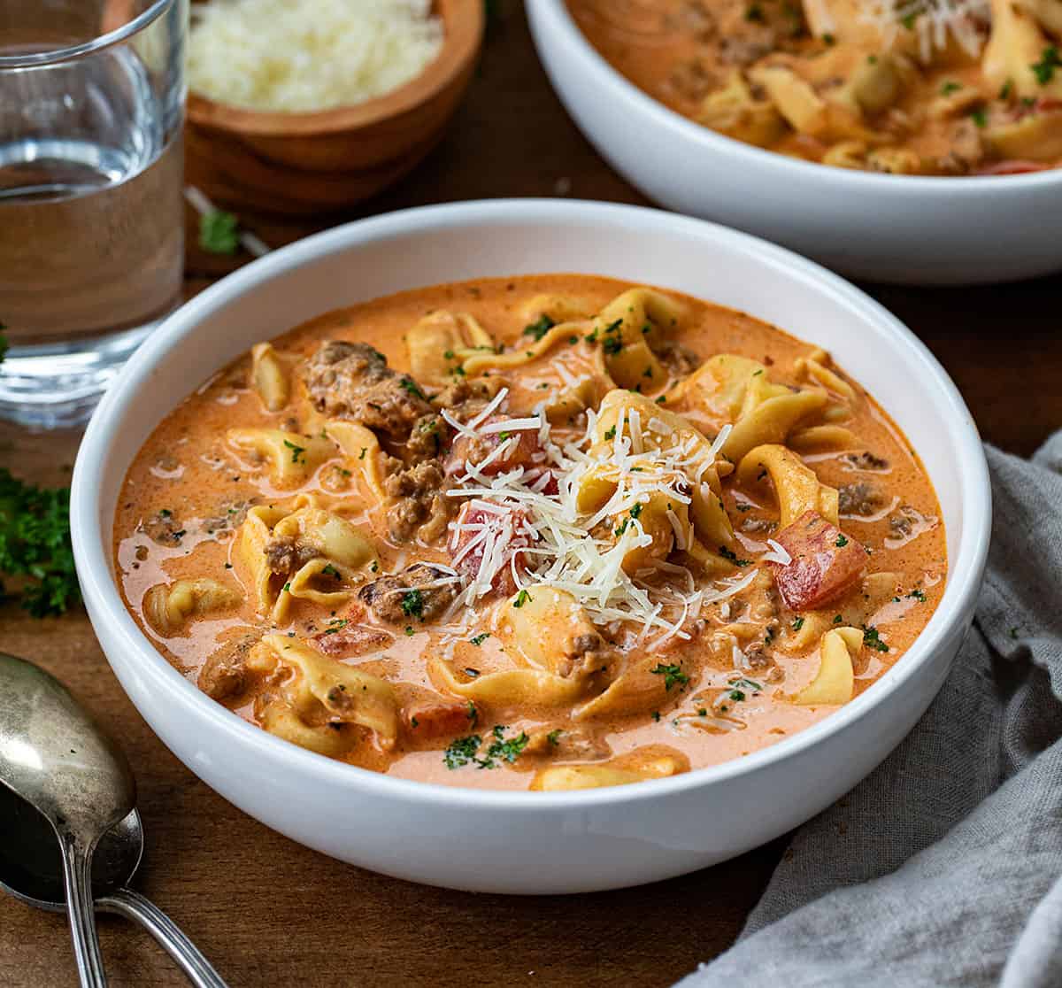 Bowls of Sausage Tortellini Soup on a wooden table with water glass and spoons nearby.