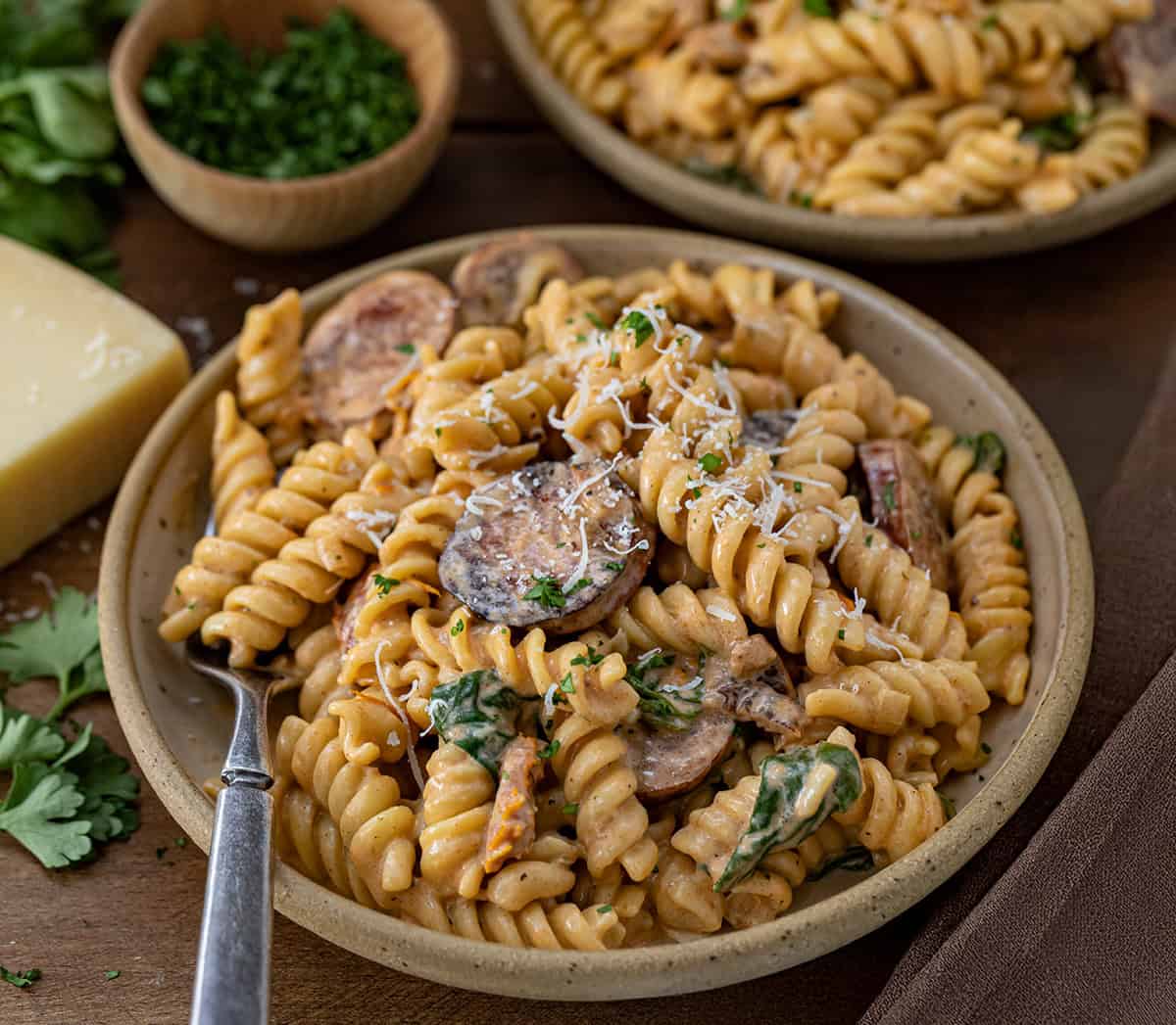 Plates of Creamy Andouille Sausage Pasta with a fork on a wooden table.