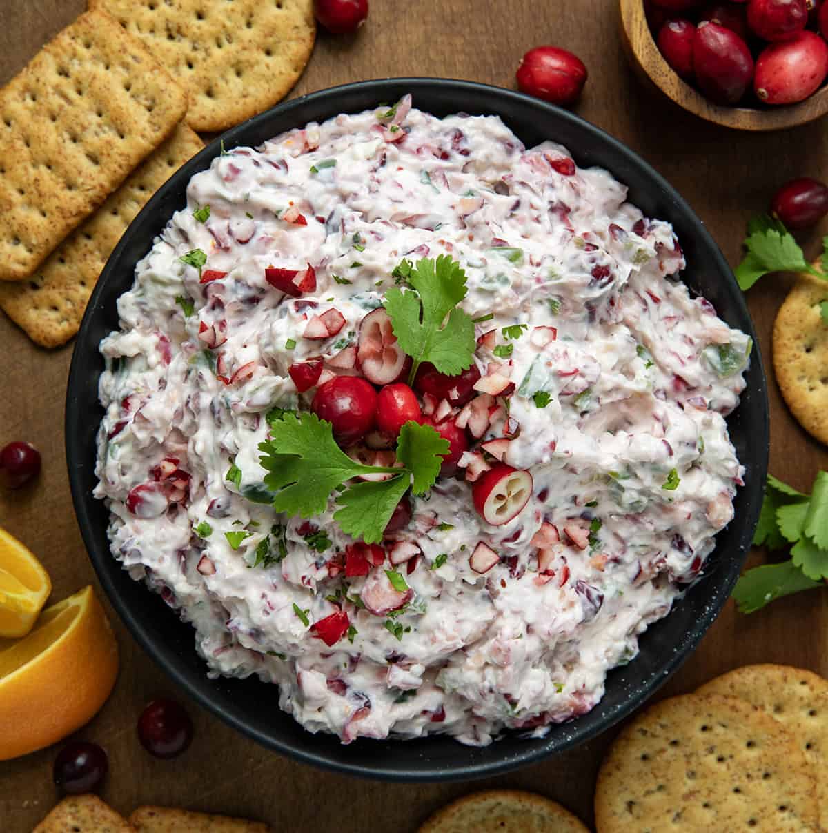 Black bowl of Creamy Cranberry Jalapeño Dip on a wooden table surrounded by crackers and oranges. 