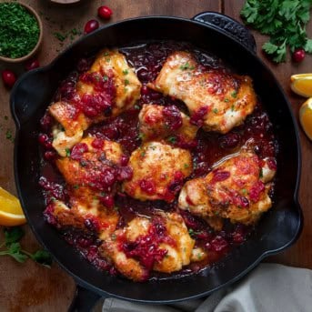 Skillet of Cranberry Orange Chicken on a wooden table with cranberries and fresh orange slices.