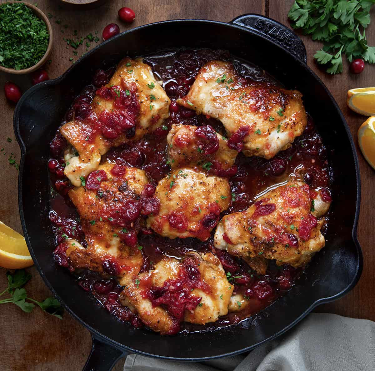 Skillet of Cranberry Orange Chicken on a wooden table with cranberries and fresh orange slices.