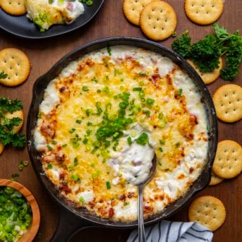 Skillet of Hot Ham and Cheese Dip with a spoon in it on a wooden table surrounded by crackers.