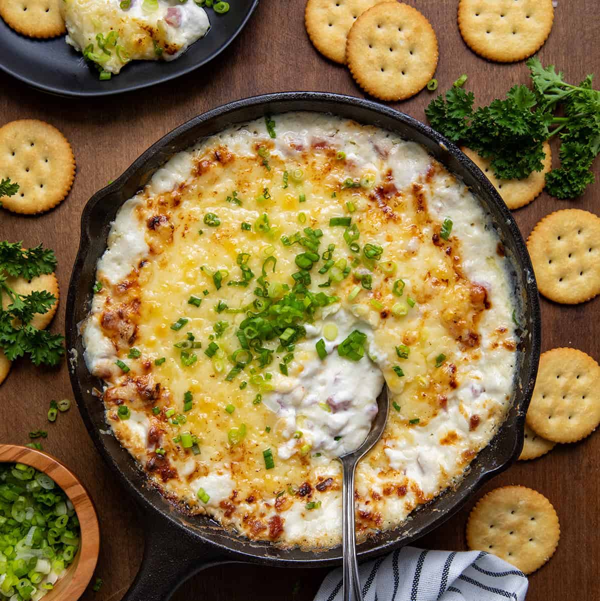 Skillet of Hot Ham and Cheese Dip with a spoon in it on a wooden table surrounded by crackers. 