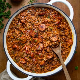 Pot of Kielbasa Cowboy Beans with a wooden spoon on a wooden table from overhead.