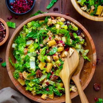 Bowl of Winter Chopped Salad with lettuce, apples, pomegranate, chicken, and feta on a wooden table from overhead with wooden spoons in the bowl.