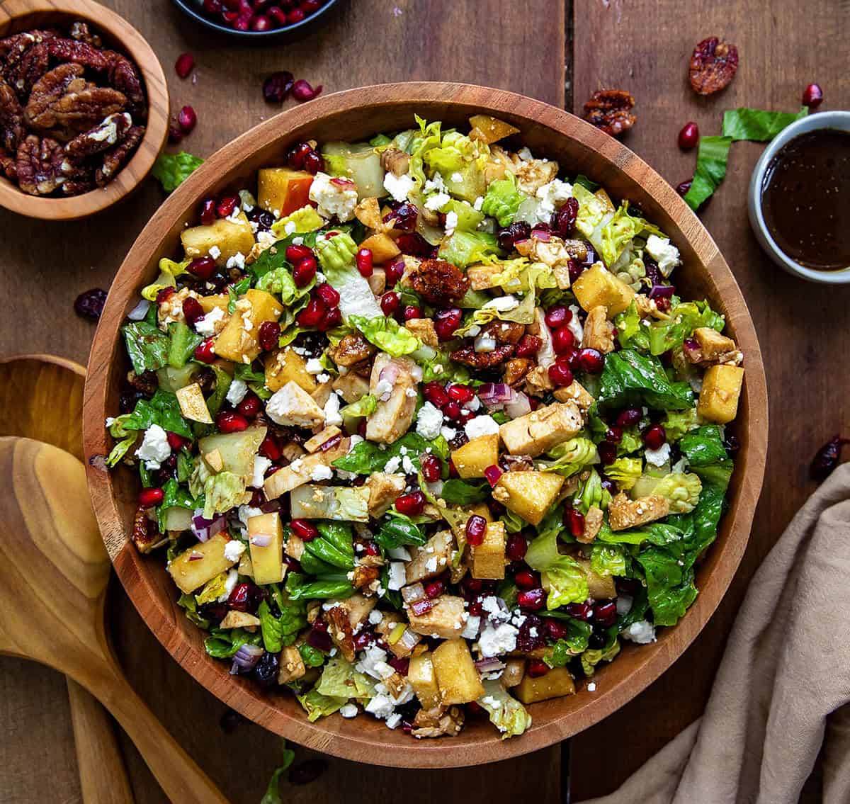 Wooden bowl of Winter Chopped Salad with lettuce, apples, pomegranate, chicken, and feta on a wooden table. 