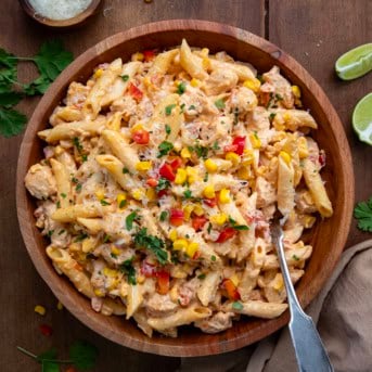 Wooden bowl filled with Sriracha Chicken Pasta Salad with a spoon in it on a wooden table with limes and freshly grated Parmesan.