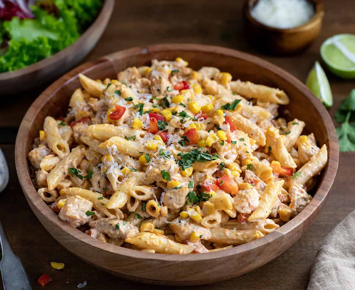 Close up of a bowl of Sriracha Chicken Pasta Salad on a wooden table with salad and Parmesan.