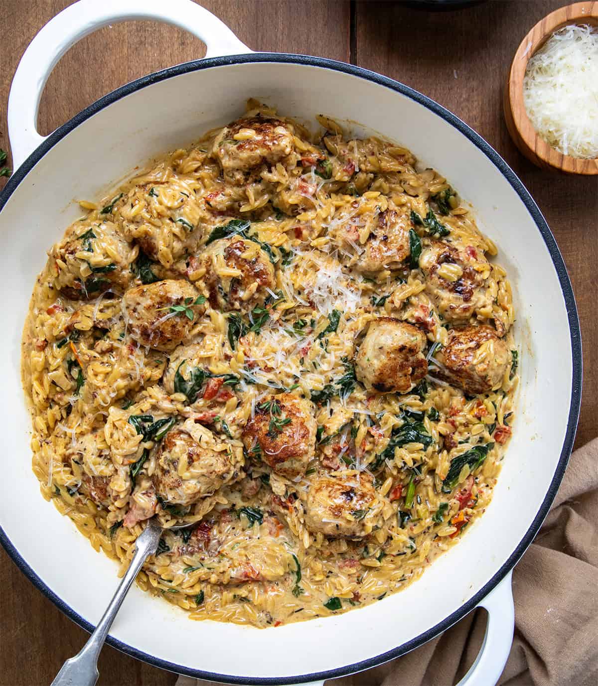 Skillet of Tuscan Chicken Meatball Orzo with a spoon in it on a wooden table from overhead.