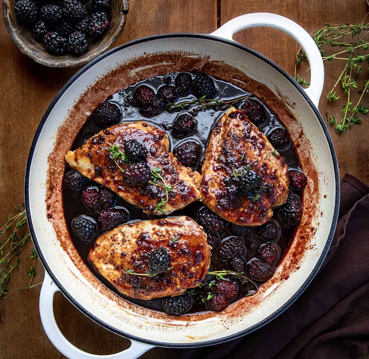 Skillet of Bourbon Blackberry Chicken on a wooden table from overhead with thyme and fresh blackberries.