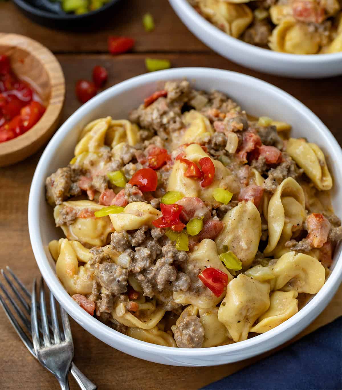 White bowls filled with One Pot Cheeseburger Tortellini on a wooden table with forks and chopped tomatoes.