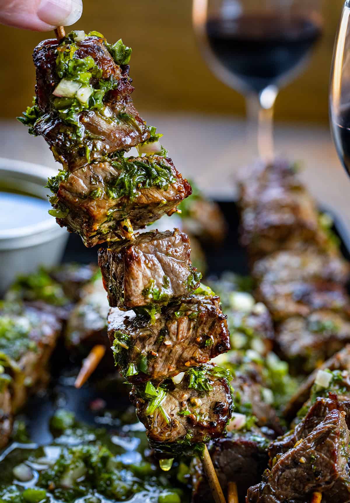 Hand holding a Chimichurri Steak Skewer above the pan with wine glasses in the background.