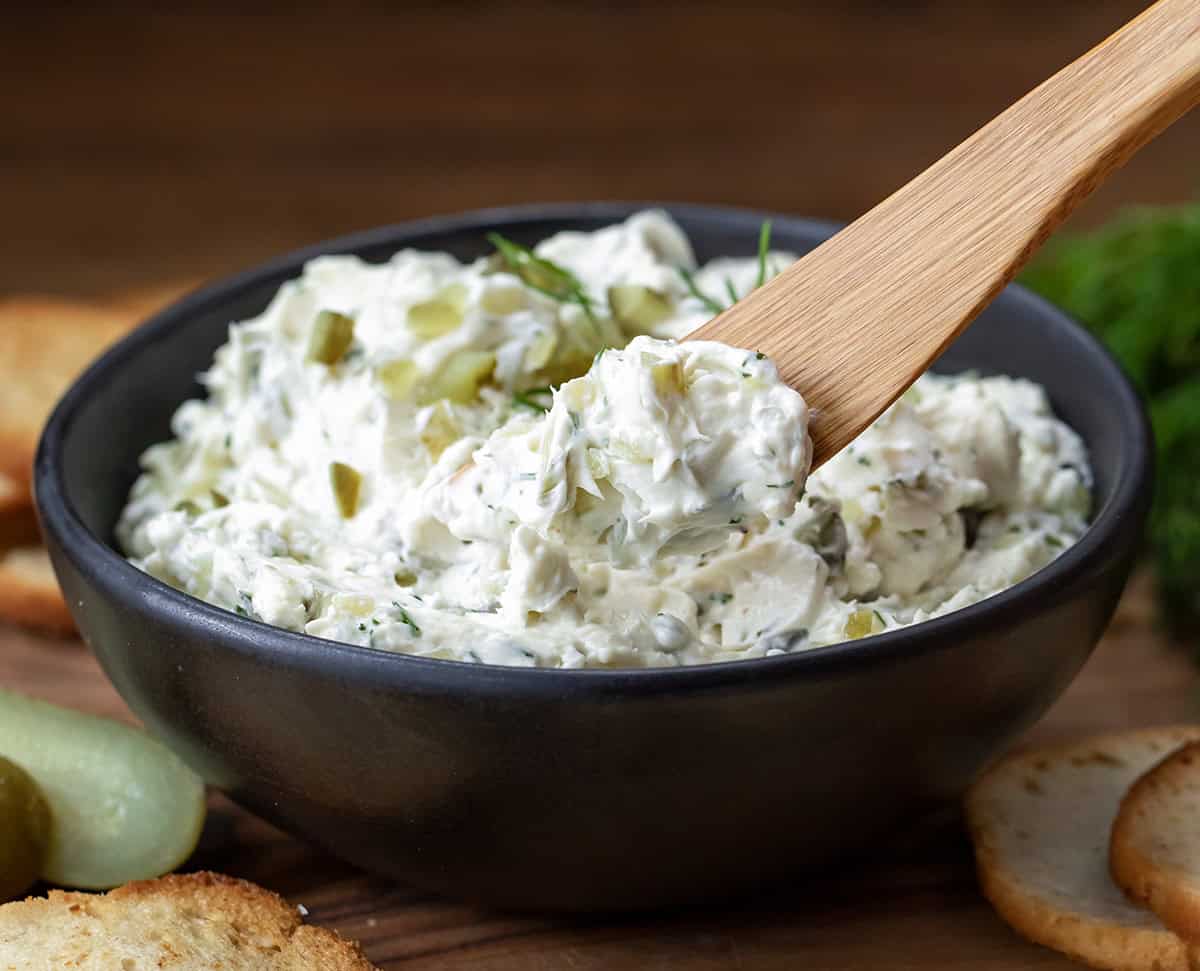Wooden knife with Dill Pickle Cream Cheese on it in front of the bowl.