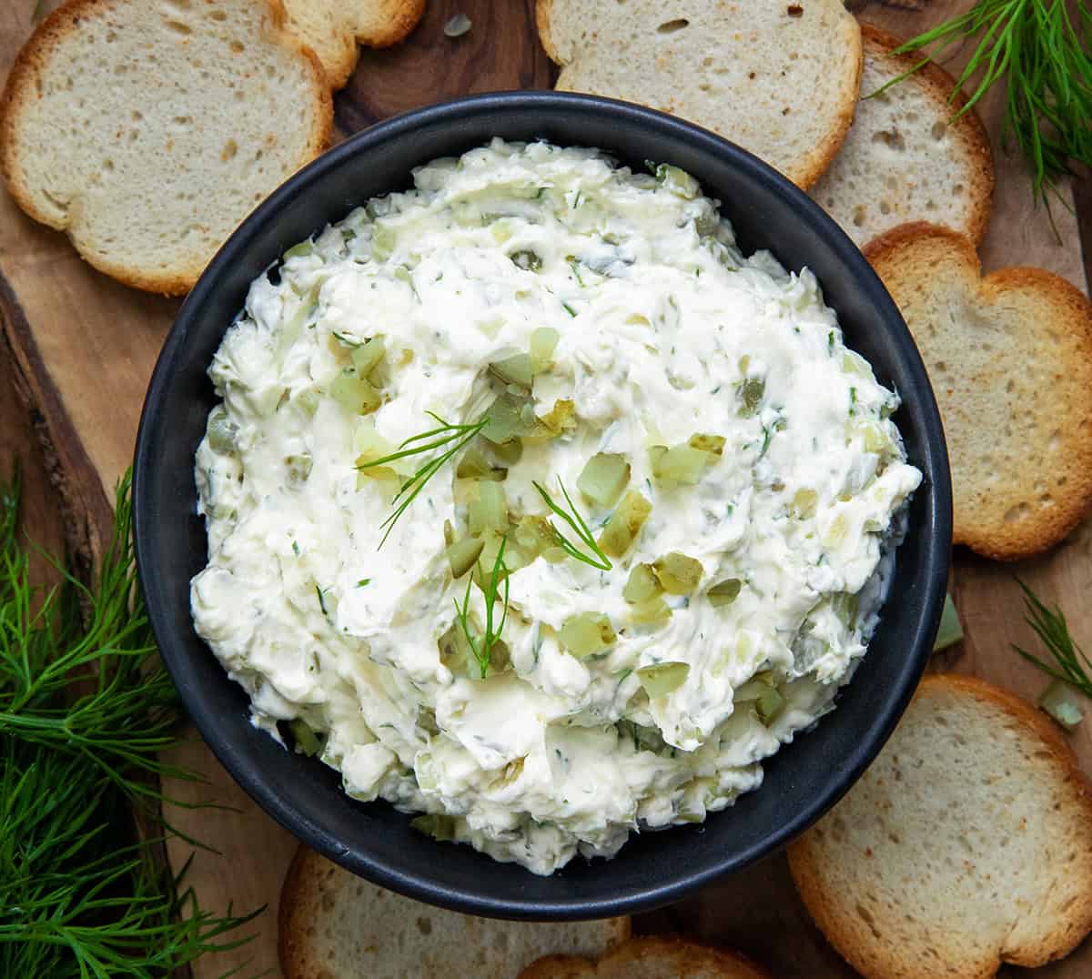 Bowl of Dill Pickle Cream Cheese on a wooden table with bagel chips all around.