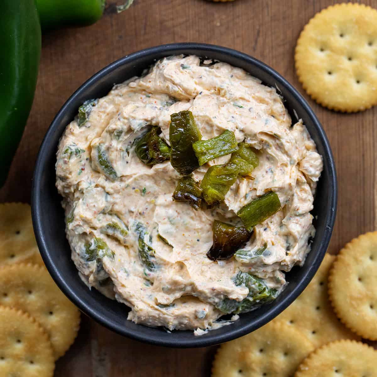 This Roasted Jalapeño Cowboy Cream Cheese is a bold and creamy dip with a spicy twist! Black bowl filled with Roasted Jalapeño Cowboy Cream Cheese with roasted jalapeños on top on a wooden table from overhead.
