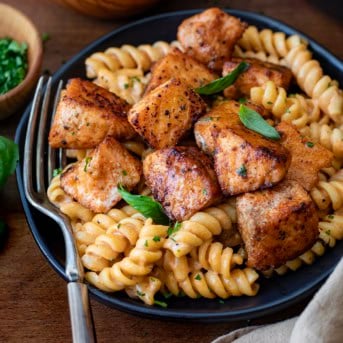 Black plate with Tomato Basil Salmon Pasta on it and a fork resting on the plate on a wooden table.