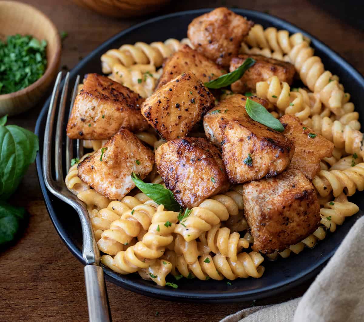 Black plate with Tomato Basil Salmon Pasta on it and a fork resting on the plate on a wooden table.