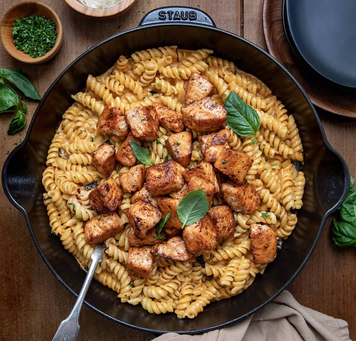 Tomato Basil Salmon Pasta is a flavorful, comforting dinner that comes together quickly (under 30 minutes!) and tastes like something you would order at a nice restaurant! Tender bites of seared salmon are served over rotini pasta tossed in a creamy tomato basil sauce made with garlic, cream, and parmesan cheese. Skillet of Tomato Basil Salmon Pasta with a spoon in it on a wooden table.