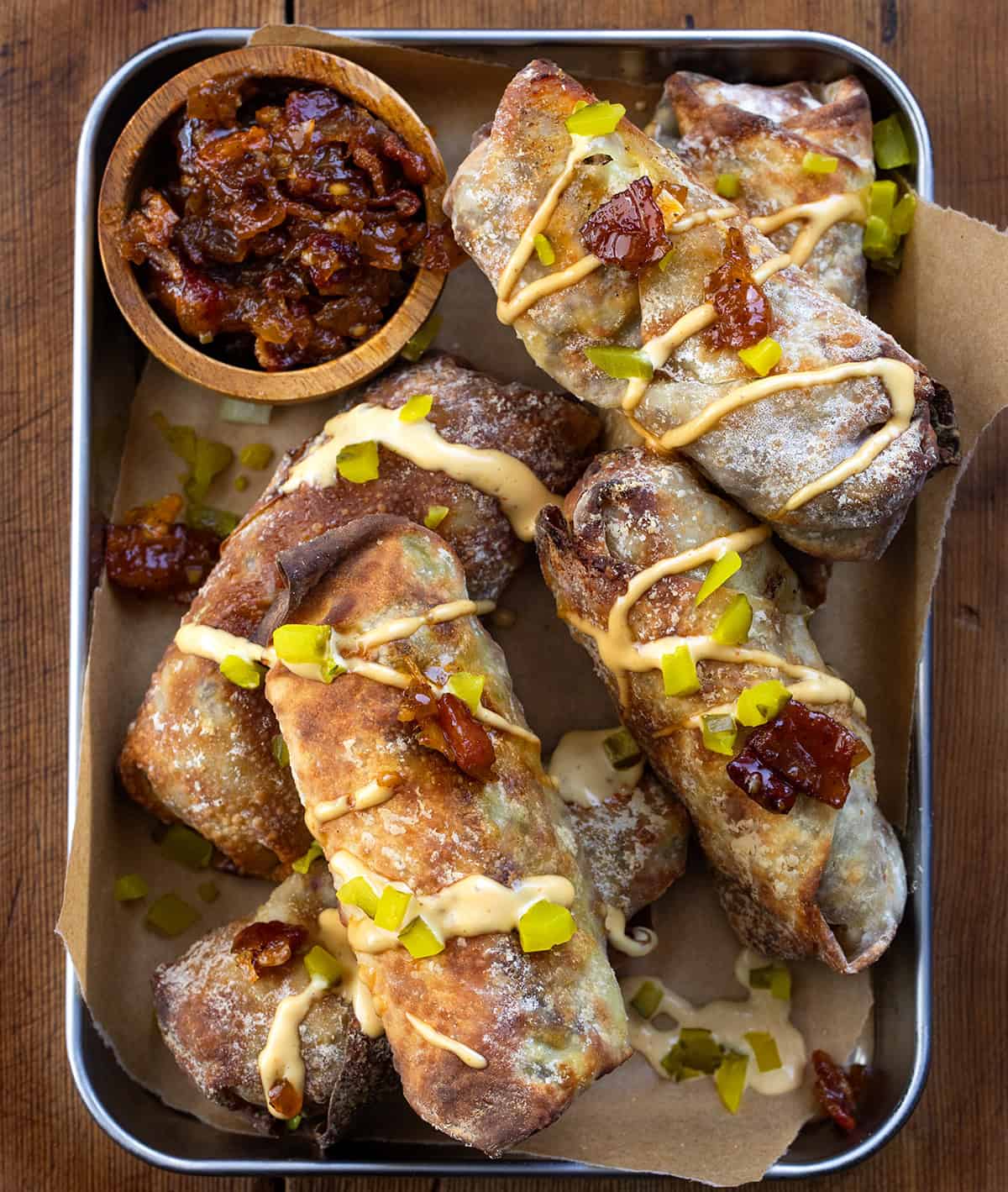 These Bacon Jam Cheeseburger Egg Rolls might be my new favorite appetizer or at least my new favorite way to enjoy a cheeseburger or egg roll! They are loaded with ground beef, melty cheddar cheese, tangy pickles, and a spoonful of my popular homemade onion bacon jam, then crisped to perfection in the air fryer. (Instructions for baking and frying in oil are also included.) Bacon Jam Cheeseburger Egg Rolls in a tray on a wooden table from overhead.