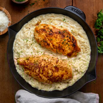 Two chicken breasts in a skillet filled with alfredo orzo on a wooden table from overhead.